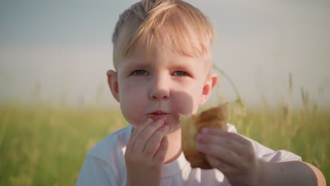 a close-up shot of a young boy wearing a white top, sitting on a grass field while eating. his expression reflects enjoyment as he savors his food in the open air