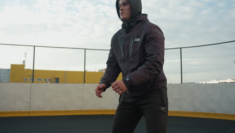 close-up of focused sportsman performing deep squats with hands clasped during workout session on outdoor sports ground, yellow industrial building and wire fence in background