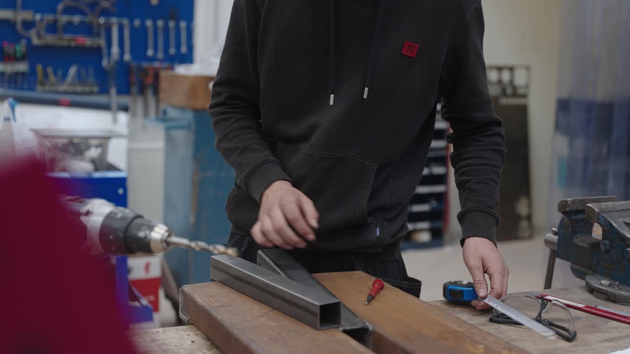 un joven aprendiz de herrero mide una pieza de metal con una cinta métrica antes de cortarla y soldarla