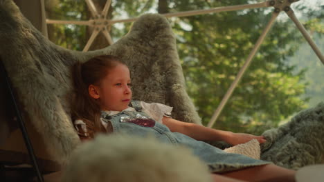 pensive preschooler girl sits in fur chair and watches nature surrounding glamping site. calm kid exploring forest outside window while sitting in cool apartment