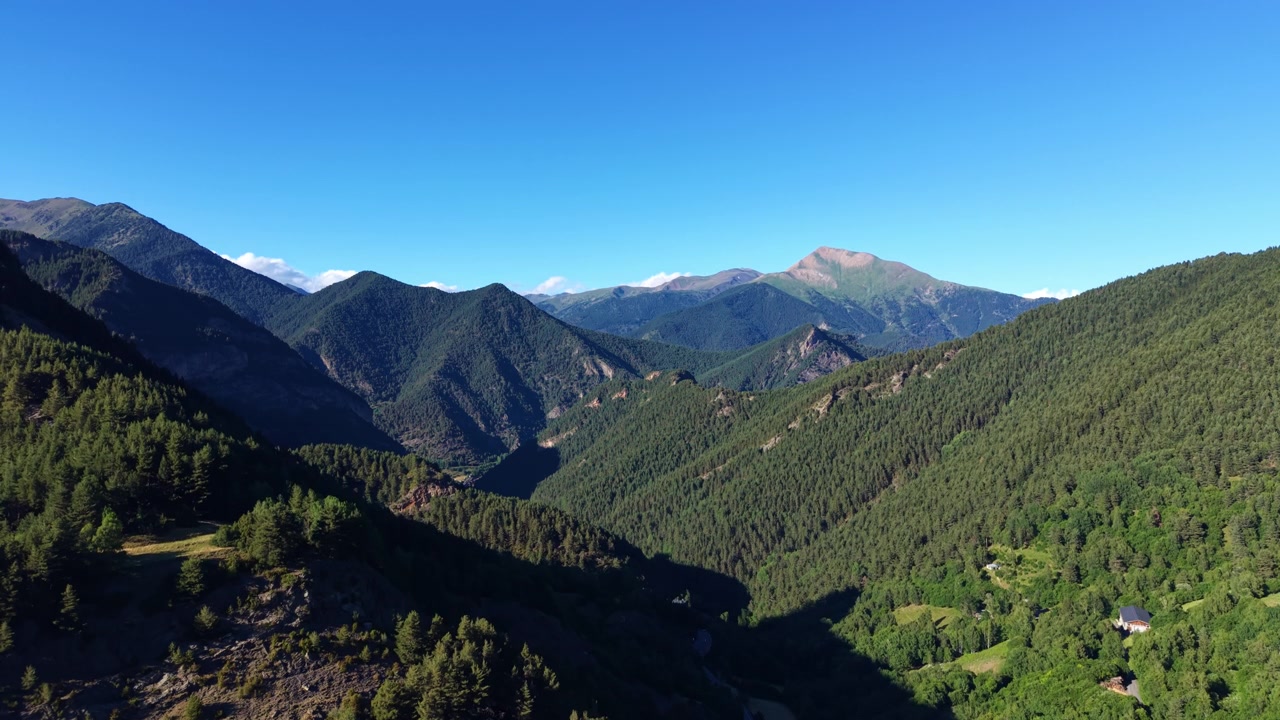 Lembah gunung berhutan lebat di Andorra di bawah langit biru cerah, dengan puncak Pyrenean berlapis membentang jauh dan sebuah bangunan kecil yang terletak di antara pepohonan.
