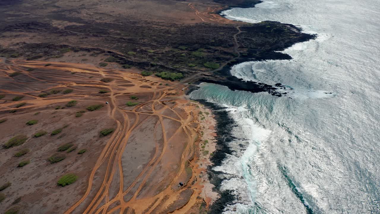 A drone sweeps along a long desert coast where off-road tracks snake to dark rocky coves and silver surf glitters under layered clouds, suggesting solitude and restless wind
