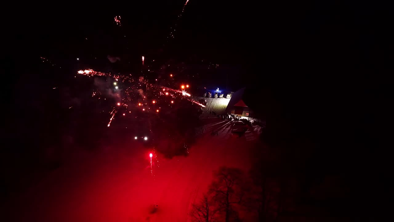 Night fireworks burst above a dark waterfront with scattered city lights, creating a festive celebration scene with bright sparks against the black sky