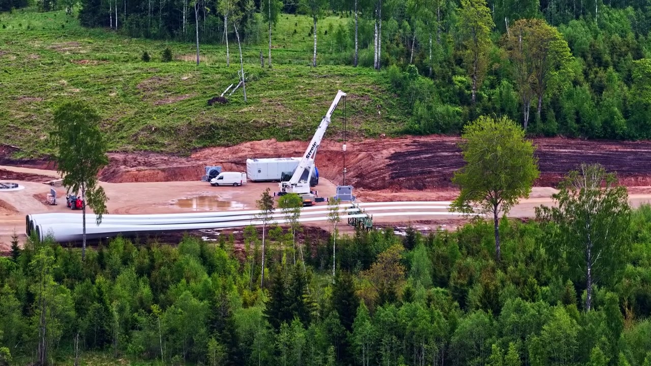An aerial shot showing the assembly process of a wind farm with turbine blades and cranes in a lush green forest clearing.