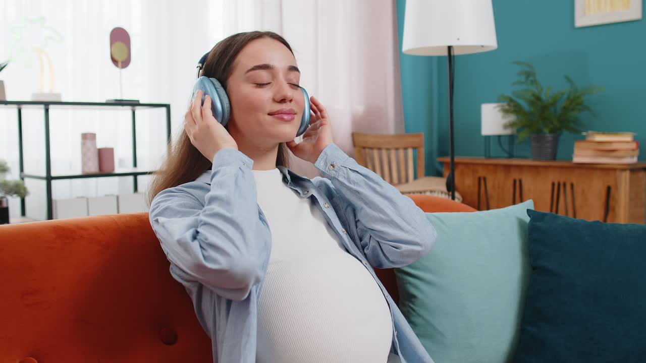 가정 소파에 앉아 음악을 듣고 아이 아기와 함께 배에 헤드폰을 착용하는 임신한 여성