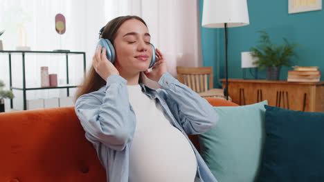 mujer embarazada sentada en el sofá de casa escuchando música poniendo auriculares en su estómago con el bebé