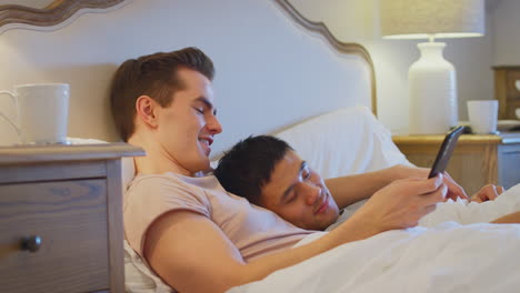 loving same sex male couple lying in bed at home taking selfie of sleeping partner on mobile phone