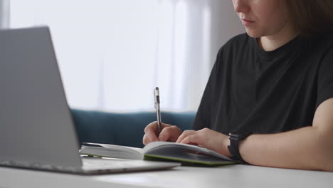 Una-Estudiante-Está-Viendo-Una-Conferencia-En-Línea-Escribiendo-Notas-En-Un-Cuaderno,-Vista-De-Cerca-Sobre-La-Mesa-Y-Aprendiendo-Cara-Desde-Casa,-Educación-Distante-Sentada-En-La-Mesa-Con-Una-Computadora-Portátil-En-El-Apartamento