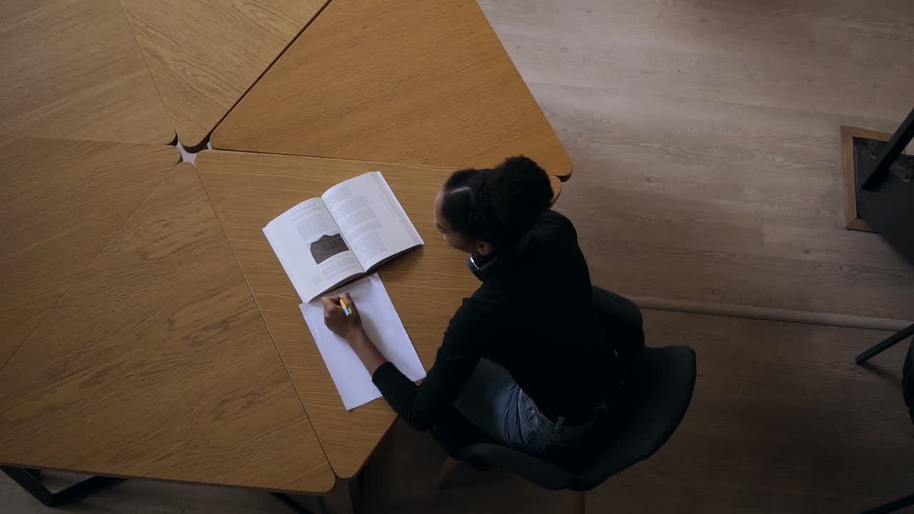 Attractive concentrated african young woman sitting by the desk and making some notes in her note book for university in the modern library, preparing for exams. Wearing black sweater and headphones. Top view footage
