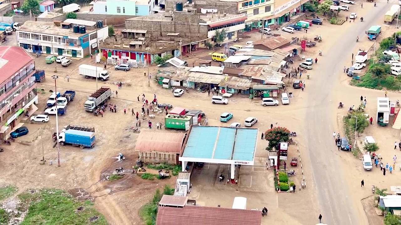 Una vista cinematografica del Kenya rurale che mostra insediamenti informali con tetti di lamiera arrugginiti, case logore, sentieri polverosi del villaggio e la vita quotidiana della comunità.