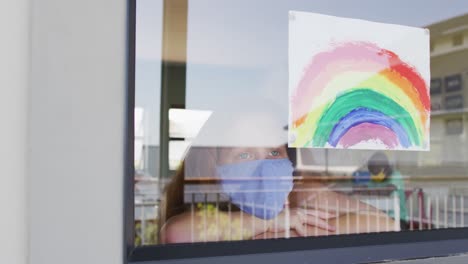 chica con máscara facial pegando pintura de arco iris a la ventana en la escuela