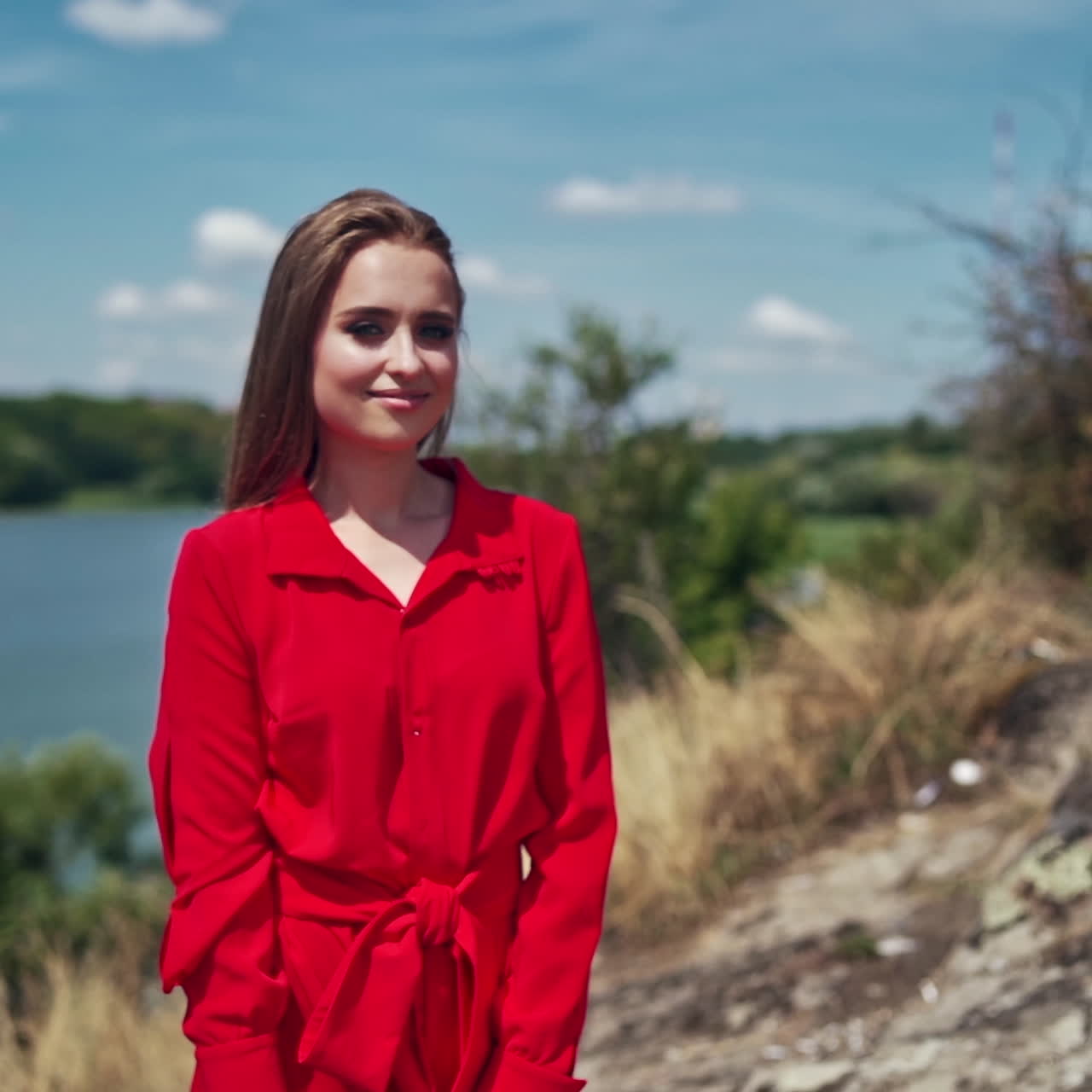 Portrait of a beautiful woman on nature background. Attractive young model in red dress with long hair standing near the river and smiling.