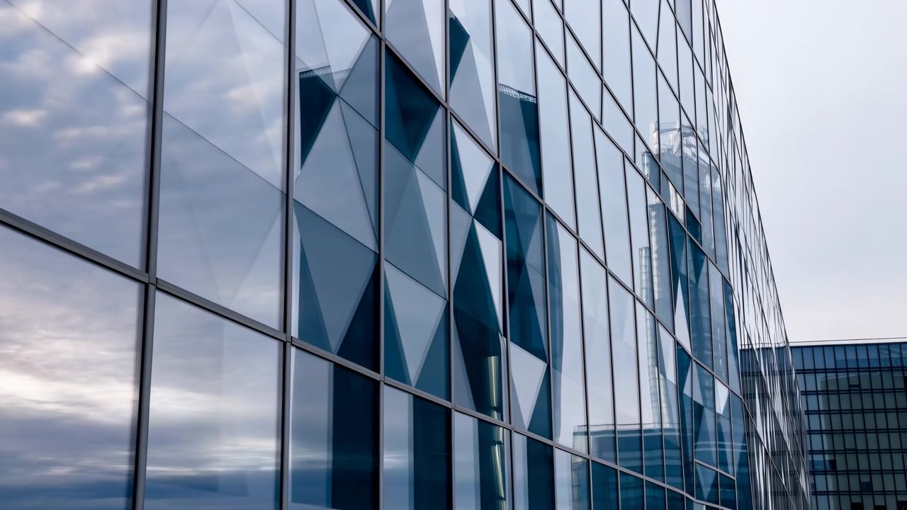 Vista a nivel de calle baja del muro cortina de un edificio de oficinas moderno que muestra acristalamiento rectangular, parteluces delgados y superficies curvas mientras las torres adyacentes reflejadas se desplazan lateralmente a través de una luz nublada fría.