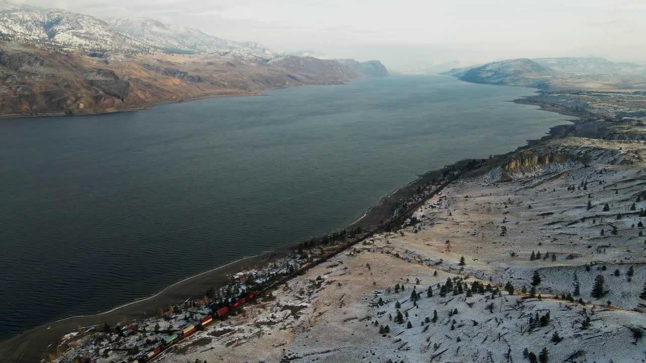 impresionante captura de un colorido tren que pasa a lo largo de la costa del lago kamloops, rodeado por un paisaje desértico montañoso parcialmente cubierto de nieve durante los primeros meses de invierno bajo la cálida luz del sol