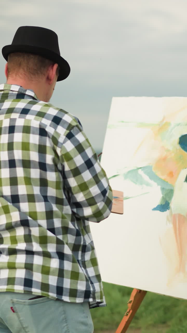 검은 모자와 체크색 셔츠를 입은 예술가가 평화로운 잔디에서 크고 다채로운 캔버스를 칠하는 동안 팔레트를 들고있는 뒷면.