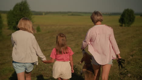 back view of mother and daughter walking closely together, with dog owner walking alongside as dog playfully reaches out for her hand, enjoying stroll in vast open field surrounded by distant trees