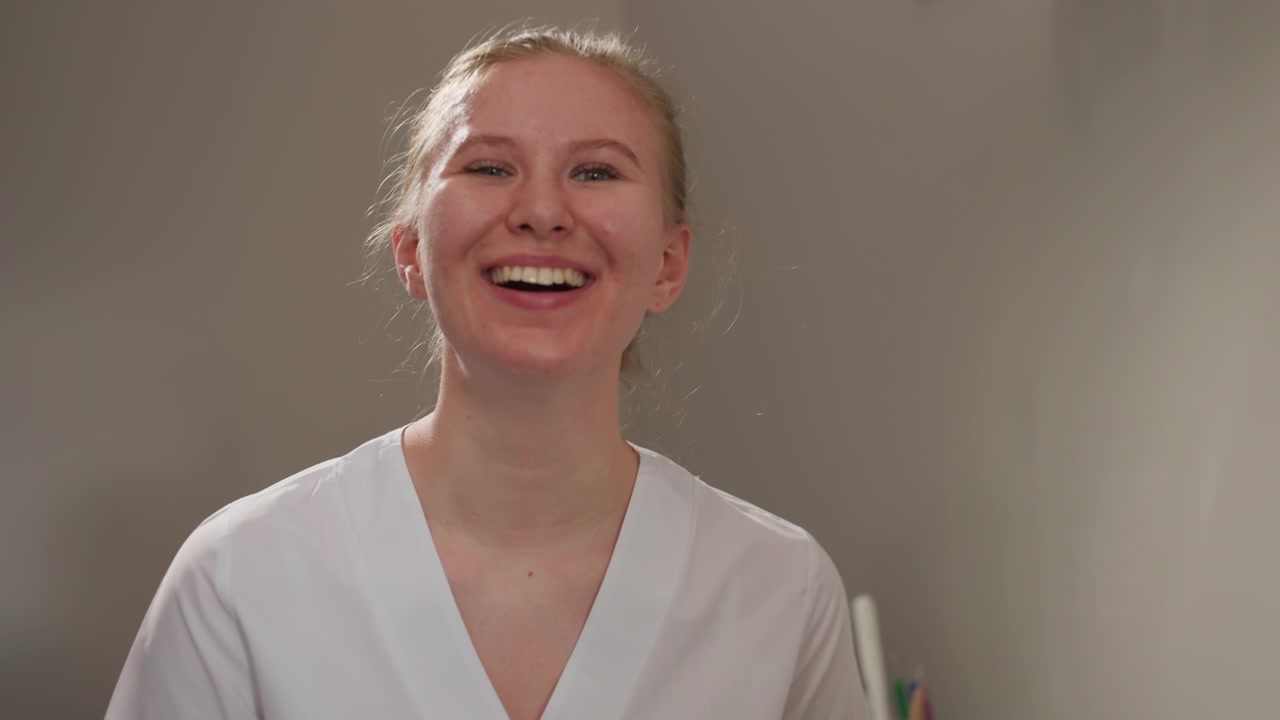 Close up of young woman in white medical uniform smiling and looking upwards. Soft lighting highlights her facial features against plain gray background with minimal camera movement