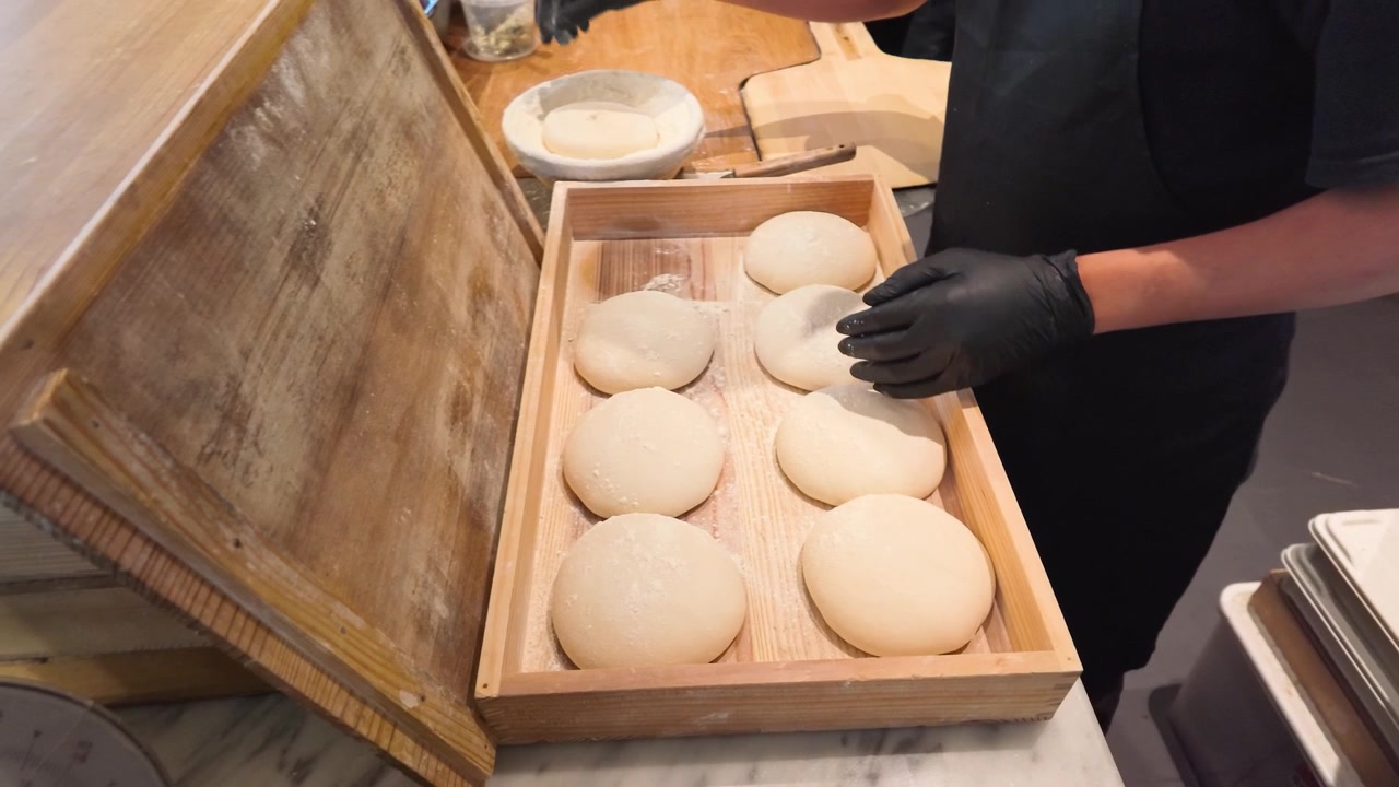 Chef profesional coloca bolas de masa de pizza cruda en una caja de fermentación de madera dentro de la cocina de un restaurante.