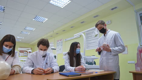 Medical students sit in row at desk and examine models of teeth carefully. Man standing at the desk puts dental instrument to the denture model.