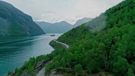 Aerial-view-following-a-coastal-road-and-a-cruise-liner-in-cloudy-Flam,-Norway