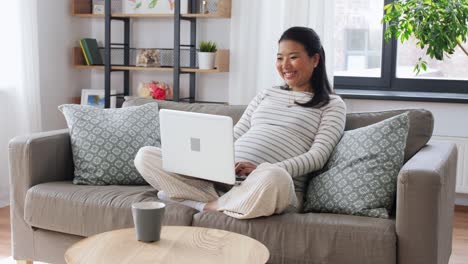 Happy-Pregnant-Asian-Woman-with-Laptop-at-Home.pregnancy,-rest,-people-and-expectation-concept--happy-smiling-pregnant-asian-woman-with-laptop-computer-sitting-on-sofa-at-home