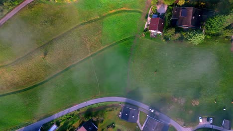 aerial view of a rural landscape with patchwork fields of green, gold, and brown, a meandering river, and several farmhouses with red roofs and tractor driving