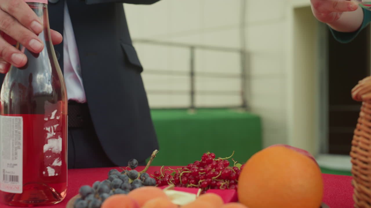 Una coppia che prepara dei calici di champagne su un tavolo da picnic, mani che stappano una bottiglia di ros accanto a un cesto di vimini, uva, arance e mirtilli rossi su una tovaglia rossa, illuminazione calda e accogliente e terrazza intima