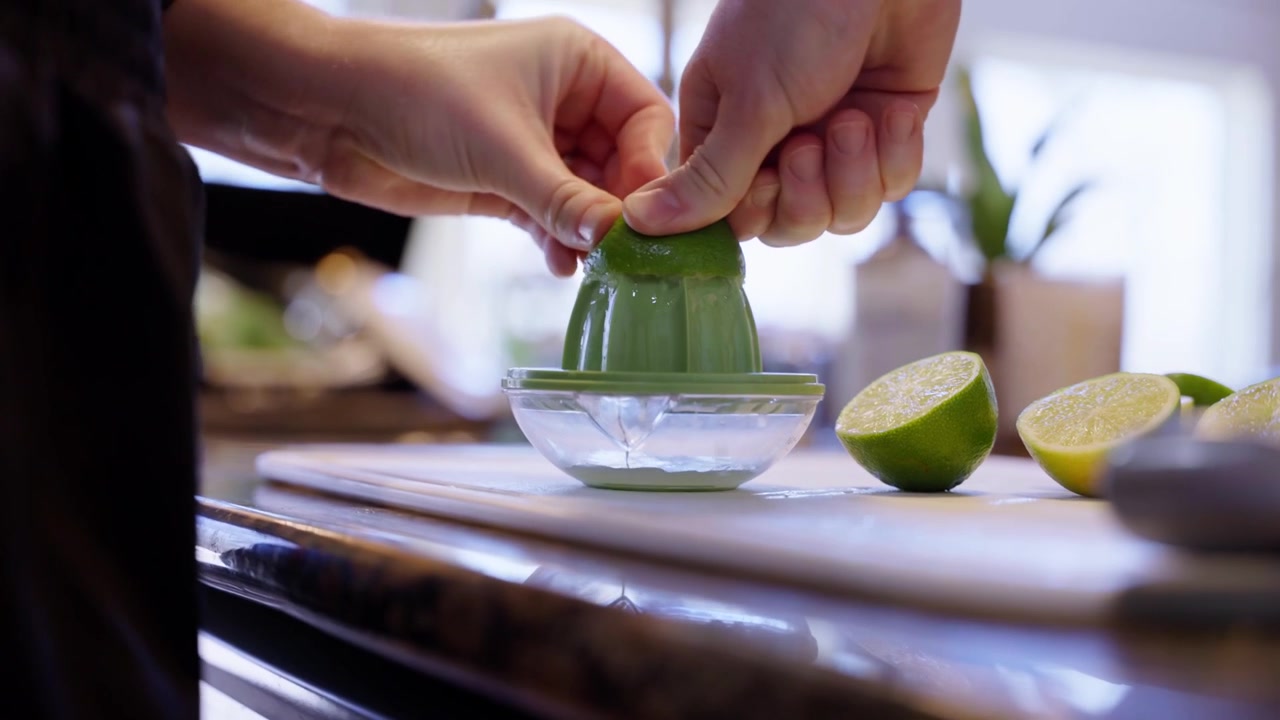 Nærbillede af en hånd, der presser en lime i en grøn manuel citruspresser, med halverede limefrugter på et køkkenbord og en lys atmosfære af hjemmelavet mad