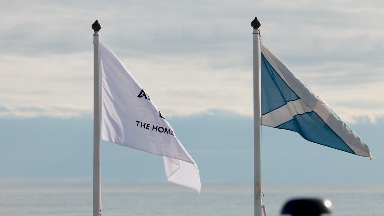 Two flags flutter vigorously on flagpoles against a cloudy sky and distant sea horizon