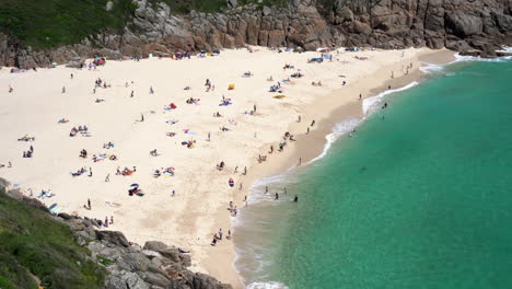 Vista-Aérea-De-La-Playa-De-Porthcurno-En-Cornwall-Llena-De-Turistas,-Nadadores,-Surfistas-Y-Familias-Divirtiéndose-En-Un-Hermoso-Día-Soleado-De-Primavera