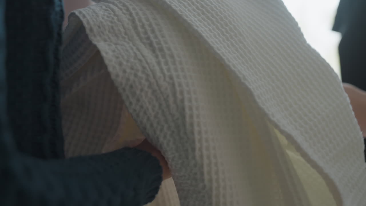 Closeup view of fair-skinned woman handing over white textured robe to another person in softly lit indoor setting, showing gesture of hospitality, comfort, and gentle interaction