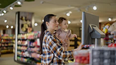 Vista-Lateral-De-Una-Madre-Con-Una-Camisa-A-Cuadros-Con-Su-Pequeña-Hija-Y-Su-Bebé-Pesando-Plátanos-En-Balanzas-Electrónicas-En-Un-Supermercado.