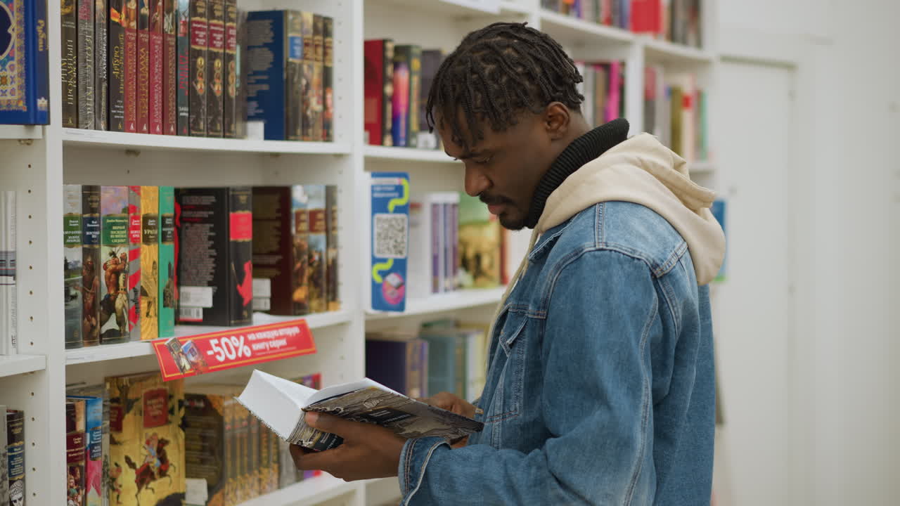 Student standing in front of library shelf, holding open book, inspecting pages with focus, surrounded by rows of colorful books, calmly exploring educational materials in modern study space