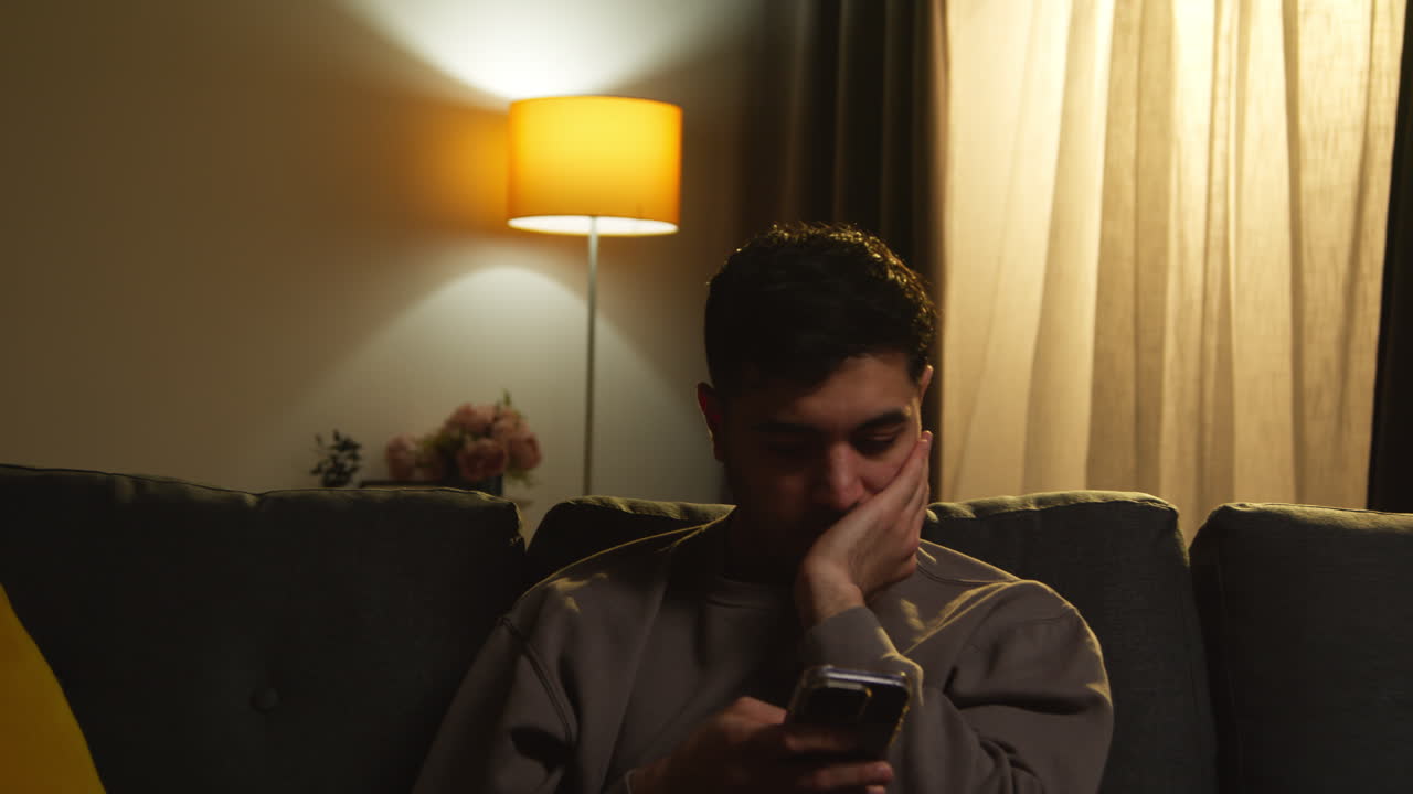 Young Man Spending Evening At Home Sitting On Sofa With Mobile Phone Scrolling Through Internet Or Social Media 3