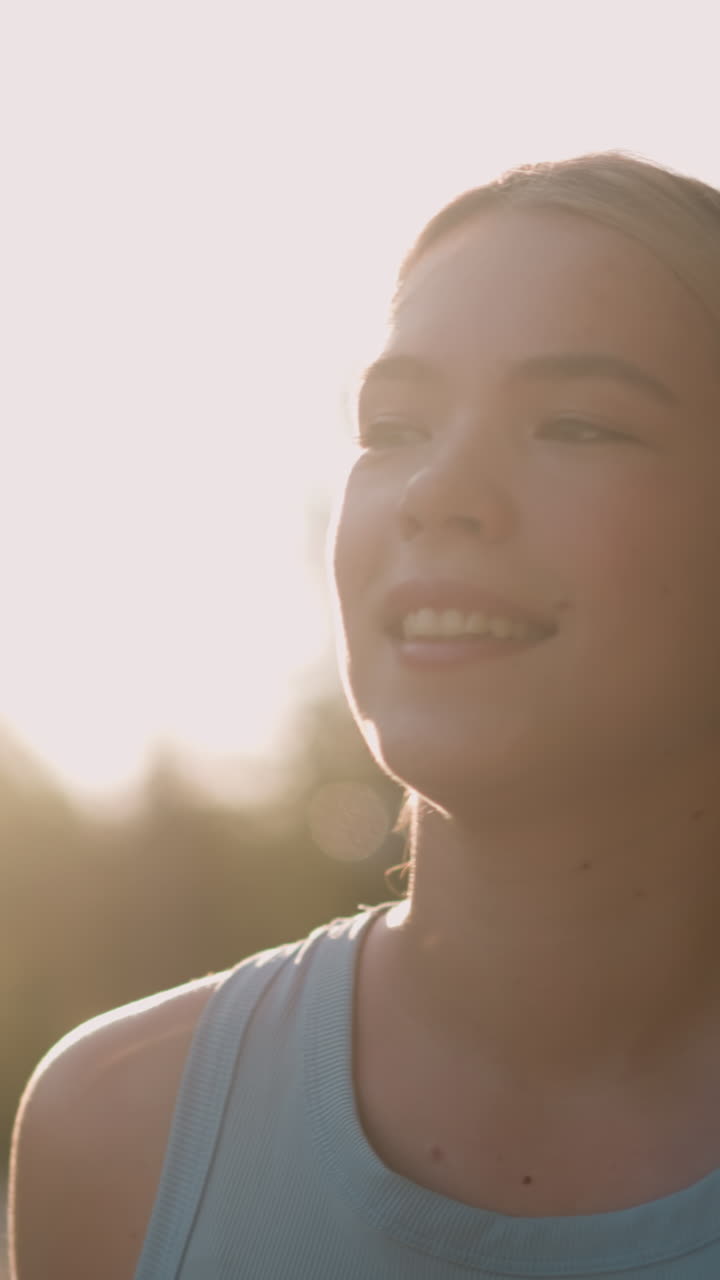 Close-up of young lady smiling with sun glowing around her face, creating a warm, radiant backlit effect, she appears serene and joyful, hair tied back, with blurred trees in background