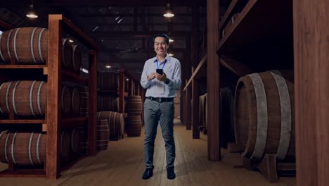 full body of an asian male professional worker standing with his smartphone in cellar of winery, he is looking at the camera with a smile