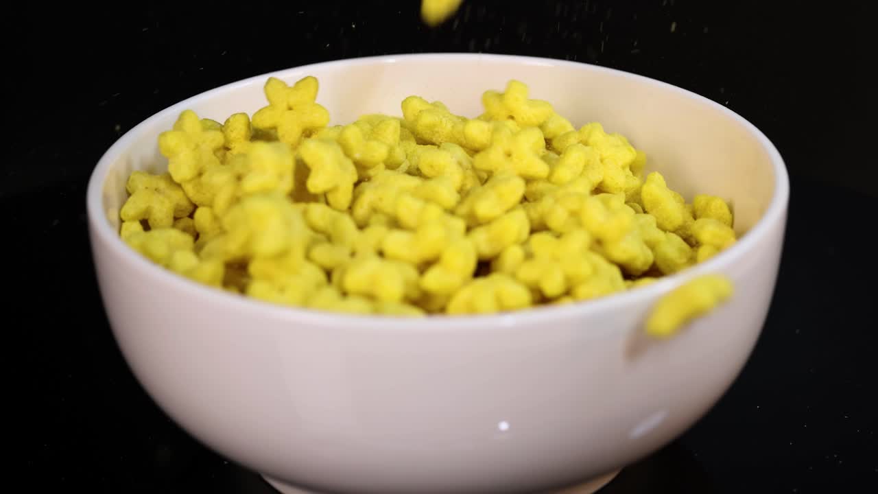Yellow cereal pieces cascade into a white bowl against a dark background, captured in slow motion with clear lighting
