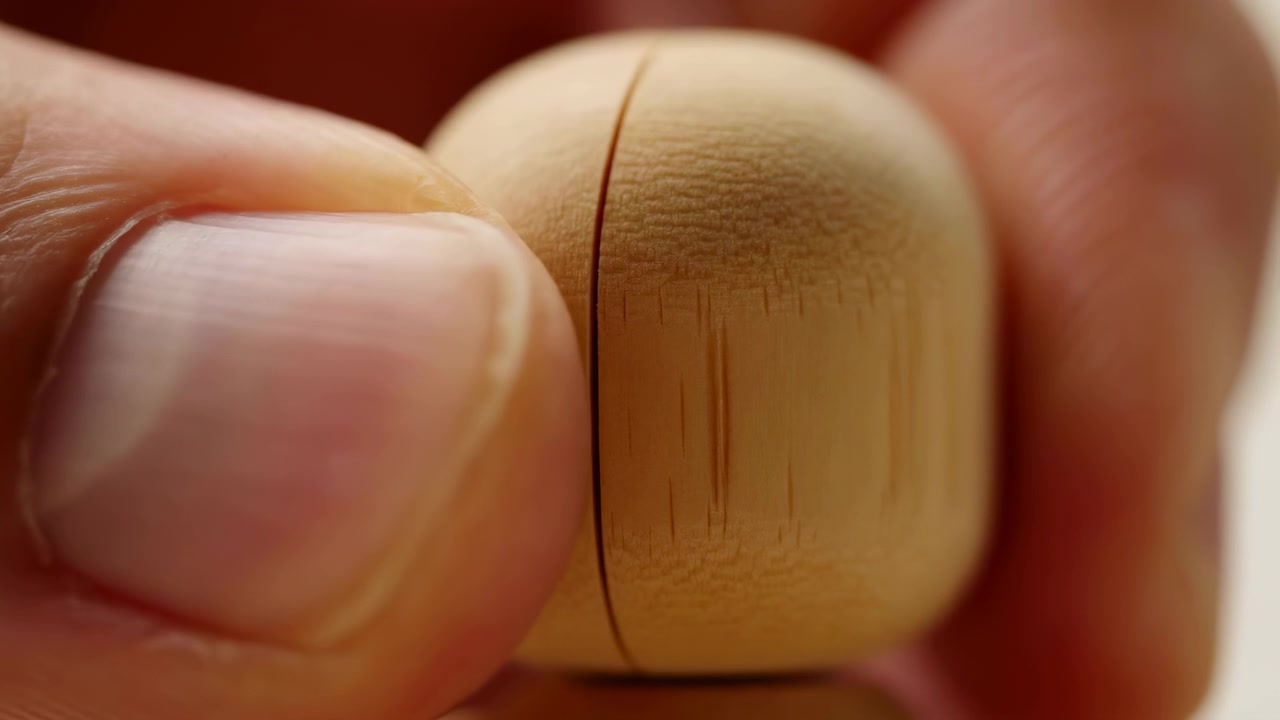 Extreme macro view shows human fingertips holding small rounded wooden object with visible grain, fine wear, warm tones, soft blur, and slight seam suggesting gentle opening in calm studio light