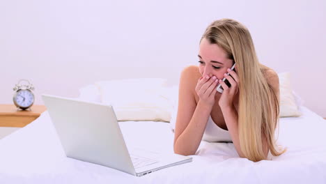 happy gorgeous blonde lying on bed phoning and using laptop