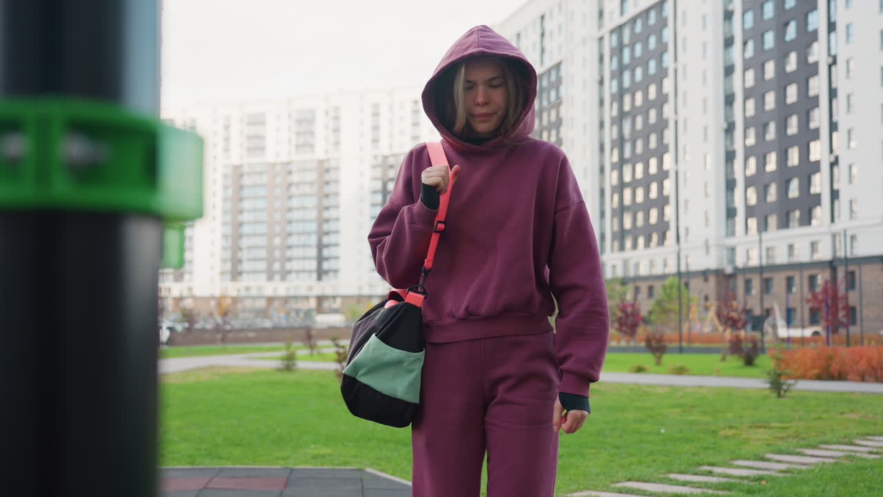 Woman walks towards exercise bar holding gym bag strap at corner of equipment under clear sky in urban outdoor fitness park near modern apartment complex, dressed in hoodie and sneakers
