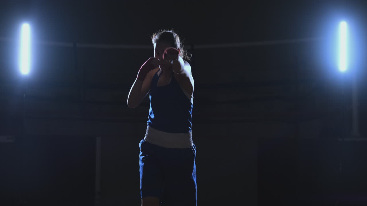 A beautiful sports boxer woman in red bandages on her hands and a blue t-shirt is fighting with a shadow practicing the speed and technique of punches. Camera movement side View. Steadicam shot. Preparing for self-defense