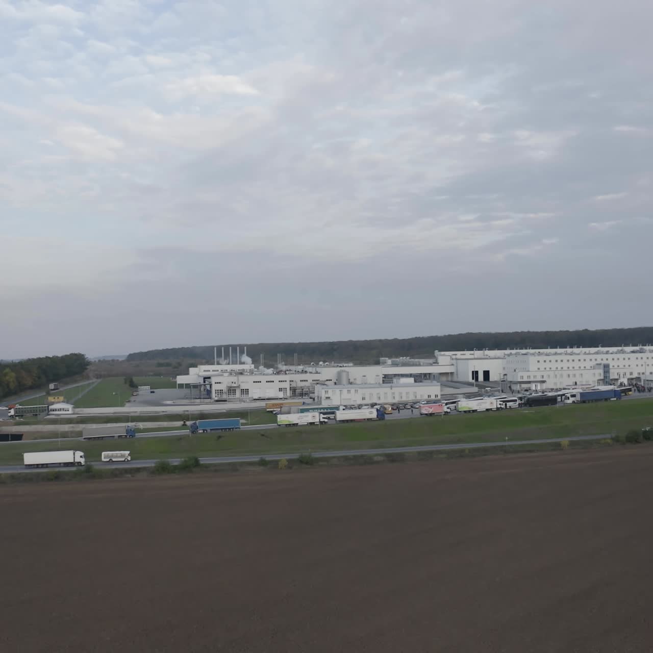 Industry among fields. Modern industrial zone in nature. Large factory with parking places with cars and trucks. Trucks moving on the road. View from the air.