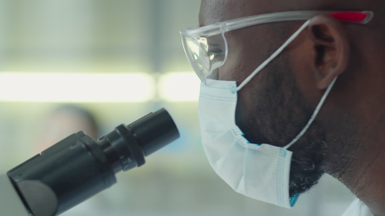 Scienziato afroamericano con maschera che lavora con un microscopio