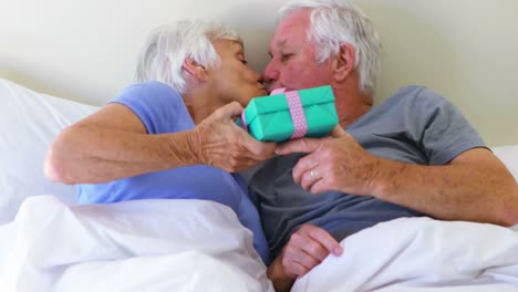 Senior-man-giving-surprise-gift-to-woman-on-bed-in-bedroom