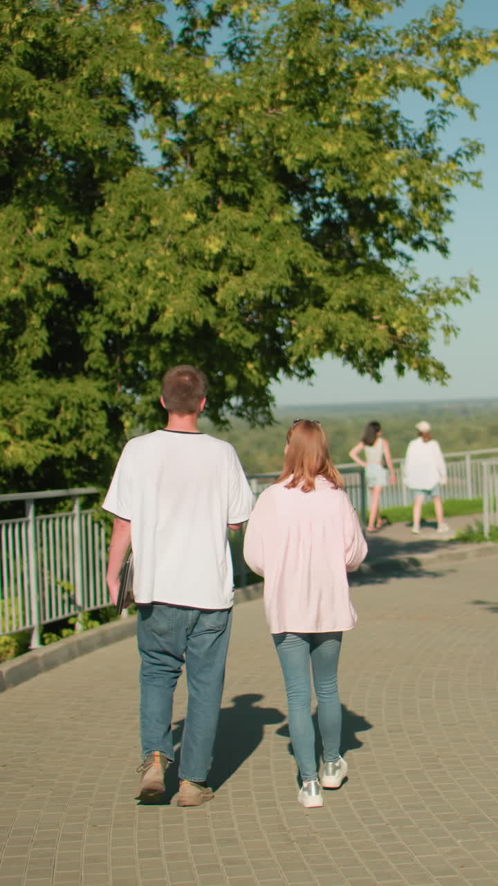 dama de camiseta rosa y vaqueros caminando junto a su amiga sosteniendo una computadora portátil en la mano izquierda cerca del riel de hierro en un camino pavimentado soleado con vegetación y bancos, dos mujeres caminando adelante, enmarcadas por árboles