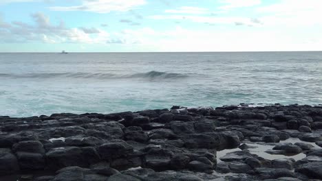 HD-Zeitlupen-Hawaii-Kauai-Boot-In-Der-Ferne-Links-Im-Bild,-Das-Von-Links-Nach-Rechts-Geht,-Mit-Meereswellen,-Die-Sich-Auf-Die-Kamera-Zubewegen,-Mit-Lavasteinen-Im-Vordergrund-Und-Teilweise-Bewölktem-Himmel