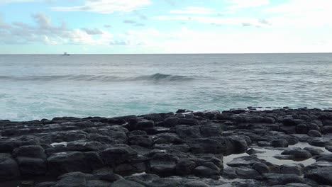 HD-Slow-motion-Hawaii-Kauai-boat-in-distance-in-left-of-frame-going-left-to-right-with-ocean-waves-building-toward-camera-with-lava-rocks-in-foreground-with-partly-cloudy-sky