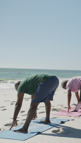 vertical video of senior african american couple doing yoga at beach, in slow motion