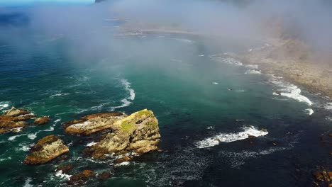 scenic aerial descending forward shot of low flying clouds above the ocean with rock formations on the coastal shore of oregon, pacific northwest