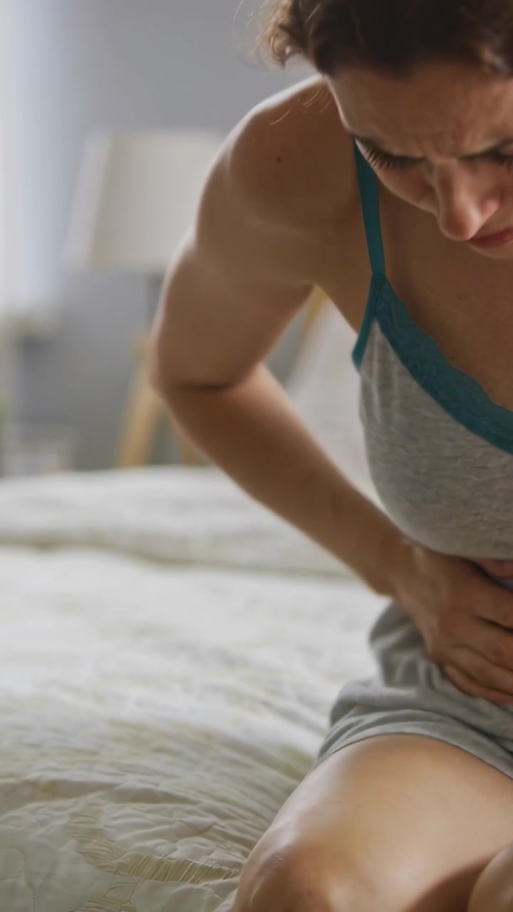 Vertical video: Wincing from cramp, woman in gray pjs, teal trim pressing abdomen in bedroom on bed